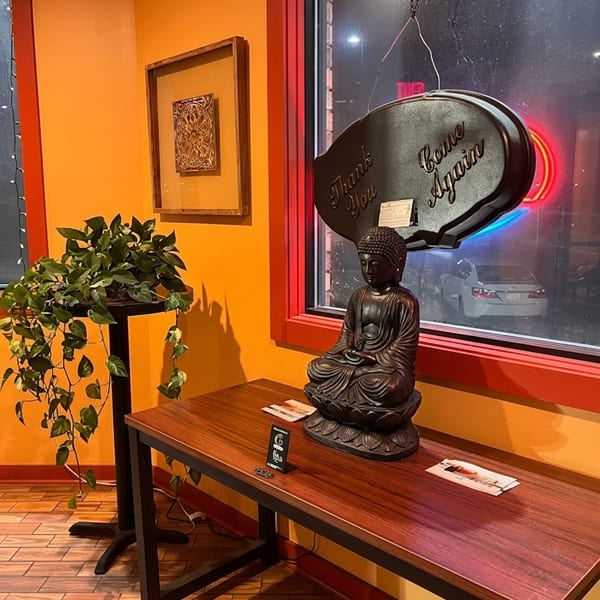Restaurant interior featuring a seated Buddha statue, hanging “Thank You, Come Again” sign, potted plant, and colorful nighttime window view.