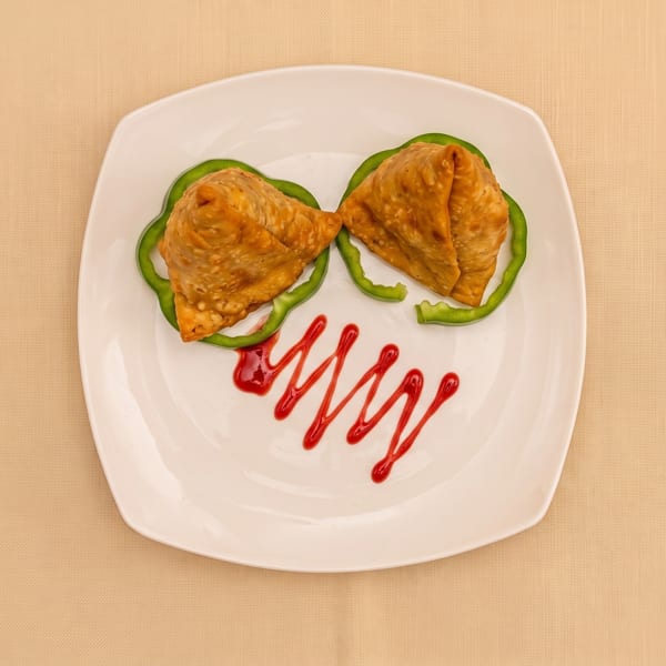Two golden fried samosas served on green pepper rings with red sauce lettering on a white plate.
