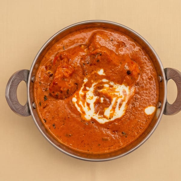 Overhead view of creamy orange Indian curry garnished with white cream in a handled metal serving bowl.
