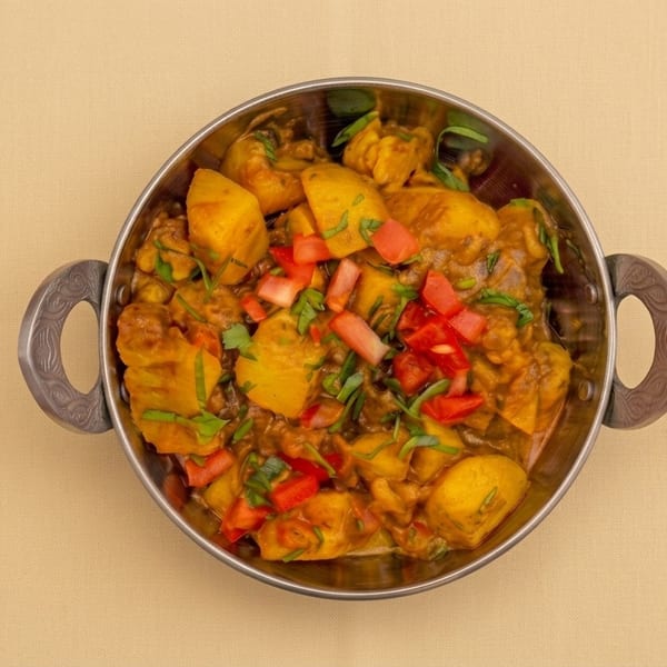 Overhead view of spiced potato curry garnished with chopped red peppers and fresh herbs in a handled metal serving bowl.