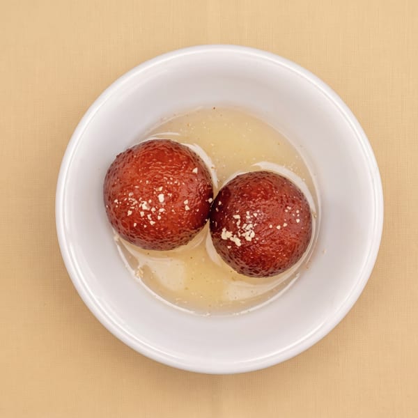 Two red-brown round desserts sprinkled with crumbs in pale sauce, served in a white bowl on a tan cloth.